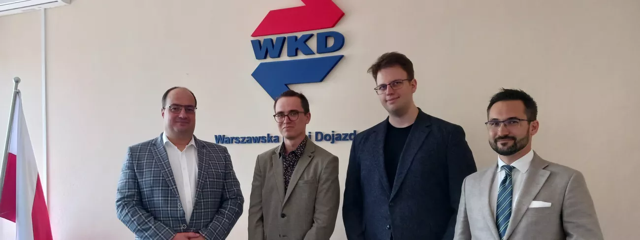 Czterech mężczyzn stojących na tle ściany z dużym czerwono-niebieskim napisem składającym się z liter WKD.
