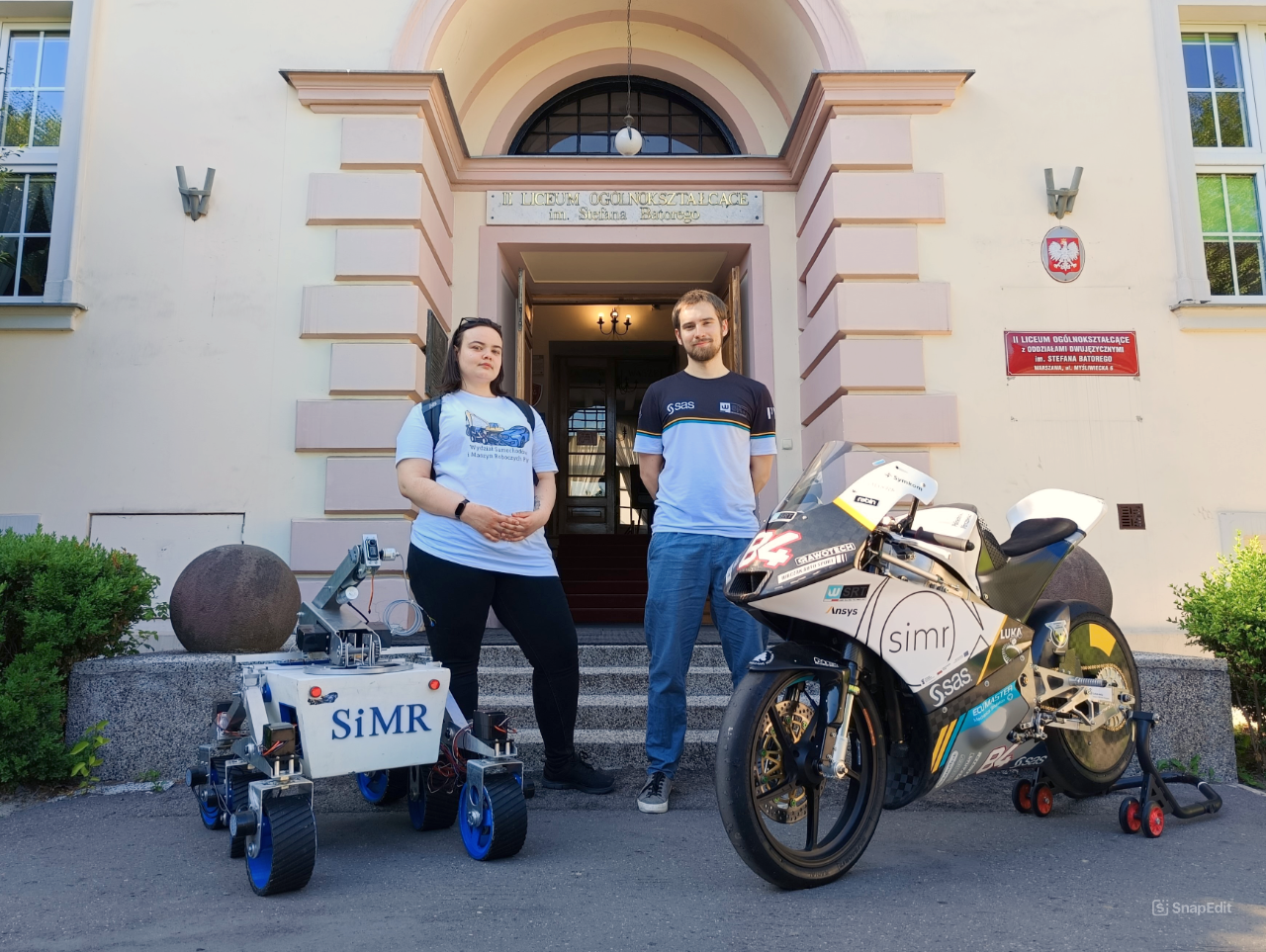 Studentka i student stoją przy łaziku marsjańskim i motocyklu wyścigowym przed wejściem do szkoły.