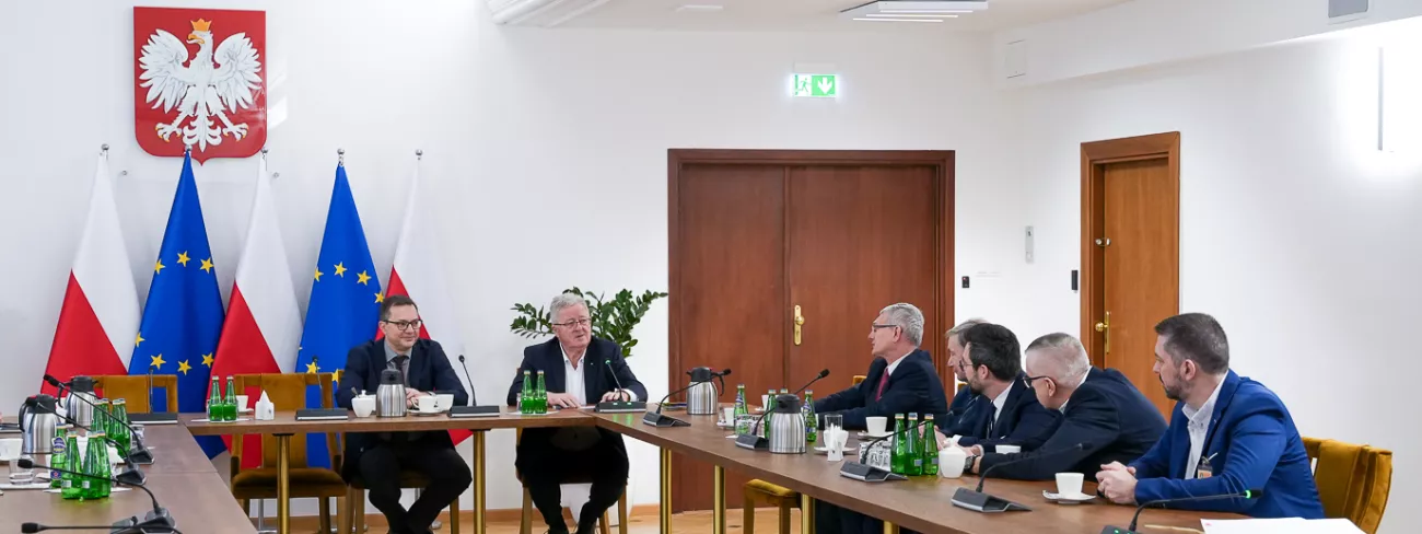 Grupa mężczyzn rozmawiających przy stole na tle flag Polski i Unii Europejskiej.