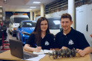 Dwie osoby siedzące za stołem warsztatowym na tle samochodów